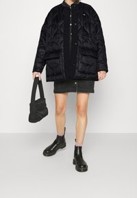 Black quilted jacket with a high collar, button closure, and two large front pockets. Paired with a black denim skirt and black ankle boots.