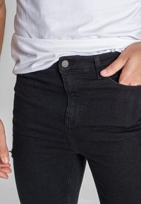 Jeans de mezclilla negra con un diseño ajustado, que cuentan con un botón de metal, bolsillos frontales y una leve textura. Llevados con una camiseta blanca.