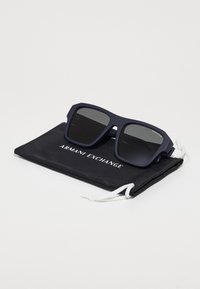 Occhiali da sole quadrati con montatura opaca blu navy e lenti scure, posizionati su un sacchetto nero con coulisse che presenta "ARMANI EXCHANGE" in testo bianco.