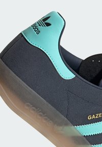 Sneaker in suede blu scuro con un inserto in plastica turchese sul tallone e tre strisce. Suola in gomma traslucida e dettaglio del logo dorato "GAZE".