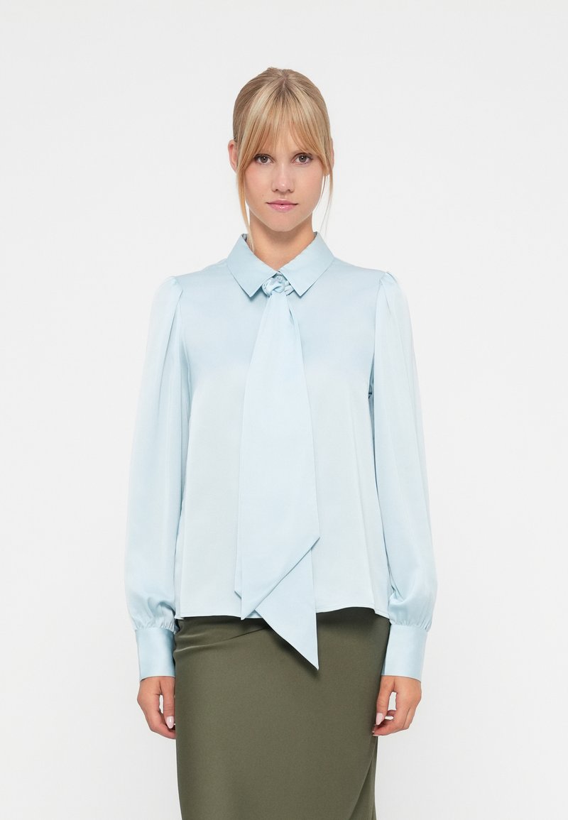 Blusa azul-clara em tecido satinado com detalhe de pescoço amarrado, mangas longas com punhos franzidos e um corte ligeiramente solto.