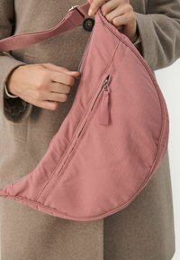 Borsa in tessuto rosa con forma curvata, dotata di chiusura a zip e una piccola tasca esterna con un pon-pon. Tracolla regolabile e materiale testurizzato.