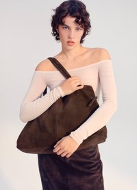 Ruda suedinė rankinė su laisva forma ir reguliuojama petneša. Matomas užtrauktukas ir siuvimo detalės. Modelis laiko ją prieš šviesų foną.