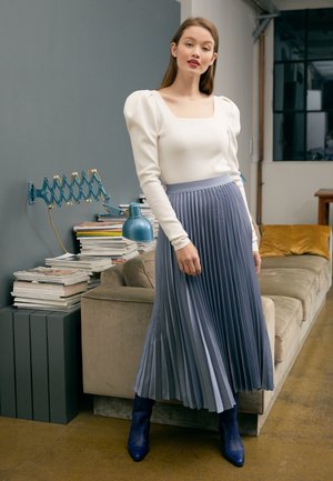 Top blanco de manga larga con mangas abullonadas, combinado con una falda midi plisada de color azul claro y botas de tobillo azules. Tejidos suaves con una textura notable.