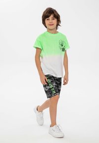T-shirt con gradient verde e bianco e stampa grafica, abbinato a pantaloni corti mimetici neri con testo verde neon, indossato con sneakers bianche.