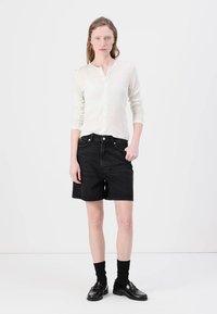 Sorte denimshorts med rett kutt, kombinert med en lys kremfarget skjorte med knapper. Modellen har på seg svarte sokker og loafers, stående mot en hvit bakgrunn.