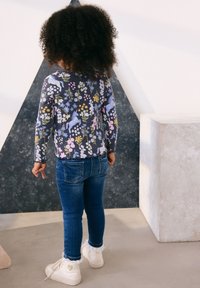 Tummansininen, pitkähihainen pusero, jossa kukkakuvio, yhdistetty sinisiin kapealahkeisiin farkkuihin ja valkoisiin nilkkureihin, joissa on pitsireunus, kuvattuna takaa.