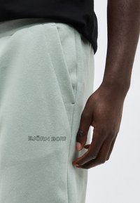 Σκούρο δέρμα χεριού που ακουμπά δίπλα σε ανοιχτοπράσινο παντελόνι φόρμας με πλαϊνή τσέπη σε γωνία και το λογότυπο "BJÖRN BORG" κοντά στην τσέπη.