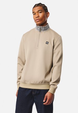 Jeune homme aux cheveux tressés portant un pull beige avec un col montant à carreaux et un jean bleu foncé sur un fond uni.