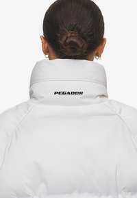 Vit pufferjacka med hög krage och synliga sömnadsdetaljer. "PEGADOR"-logotyp i svart på baksidan, slät textur, lätt och smidig design.