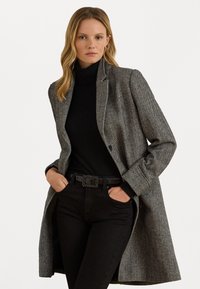Sivý kabát s herringbone vzorom, so šikmými klopami, zapínaním na jediné gombík a dlhými rukávmi, oblečený cez čierny rolák a čierne džínsy.