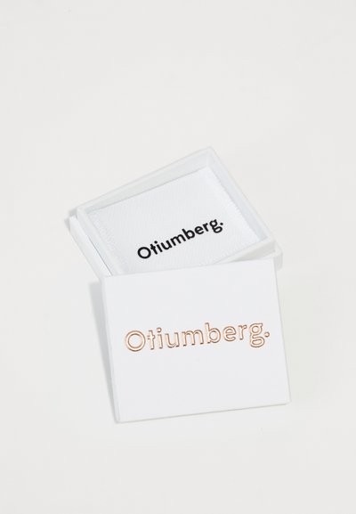 Weißer quadratischer Schmuckkasten mit einem rosefarbenen "Otiumberg."-Logo auf der Vorderseite und einem weißen Innenraum, der ein schwarzes "Otiumberg."-Etikett zeigt.