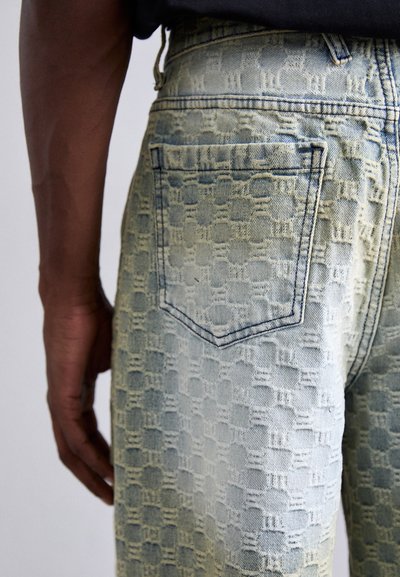 Jean en denim bleu clair avec un motif en relief. Présente une poche arrière et des coutures visibles, mettant en valeur la surface texturée.