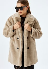 Abrigo de piel sintética beige con cuello acanalado, diseño de doble botonadura y botones negros. La modelo posa con una blusa de encaje clara y gafas de sol oscuras.