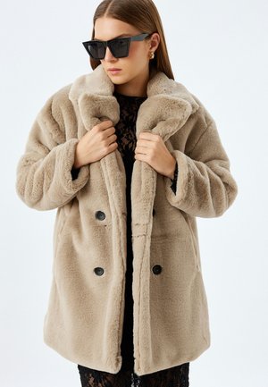 Cappotto in pelliccia sintetica beige con colletto a scollo, design doppiopetto e bottoni neri. Il modello posa con un top di pizzo chiaro e occhiali da sole scuri.