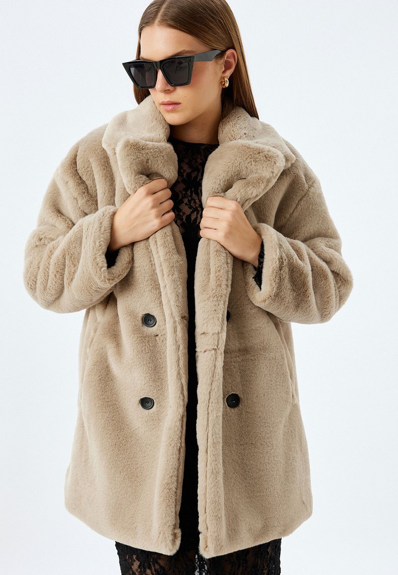 Abrigo de piel sintética beige con cuello acanalado, diseño de doble botonadura y botones negros. La modelo posa con una blusa de encaje clara y gafas de sol oscuras.