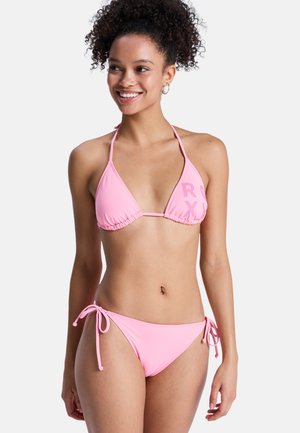 Mosolygó, göndör hajú nő, rózsaszín háromszög alakú bikiniben, oldalt megkötve, egyszerű fehér háttér előtt áll.