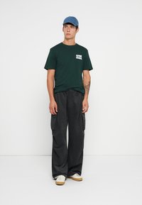 Grønn bomull T-skjorte med teksten "ALPINE GOODS", svarte vide cargo-bukser og en blå denim-kaps, iført med lyse sneakers.
