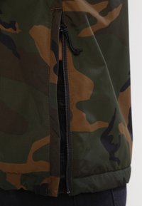 Camouflagejakke i mørke grønne og brune nuancer med lynlåslukning og elastisk kant. Materialet har en glat tekstur med vandafvisende finish.