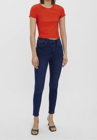 Top corto de manga corta de rayas rojas con bordes en forma de onda, combinado con unos jeans ajustados de cintura alta de color azul oscuro. La modelo lleva sandalias de tacón abierto verdes.