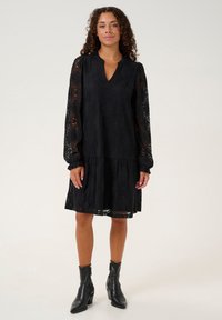 Vestido de encaje negro con mangas largas y escote en V. Cuenta con una falda en capas y patrones florales intrincados. Combinado con botines negros.