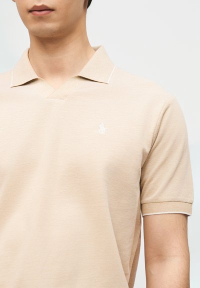 Άνδρας που φοράει μπεζ πόλο μπλουζάκι με κοντά μανίκια, γιακά και μικρό λευκό κεντητό λογότυπο στο στήθος.