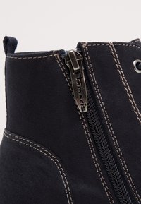 Bottine en daim noir avec une fermeture éclair latérale, des surpiqûres contrastantes et une languette à l'arrière pour un enfilage facile.