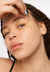 Gotero de suero transparente sostenido sobre la mejilla de un modelo, con una tapa plateada, líquido ámbar y una textura de piel visible con pecas.