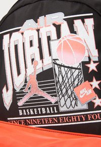Sac à dos noir arborant un grand graphique avec le texte "Air Jordan", un ballon de basket et une silhouette d'un joueur, rehaussé d'orange dans la partie inférieure.