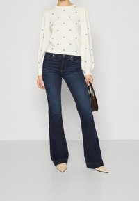 Persoon in een wit langmouwtrui met kleine blauwe bloemen, donkerblauwe wijde jeans, beige puntige schoenen en een kleine bruine handtas vasthoudend.
