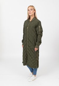 Pikowana oliwkowa długa kurtka z zamkiem z przodu, ściągaczami przy rękawach oraz kieszeniami bocznymi. Posiada złote akcesoria i luźny krój.