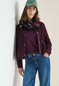 Burgundy fuzzy cardigan with snap buttons over a dark purple sweater, paired with blue denim jeans and a patterned black, red, and blue scarf.
