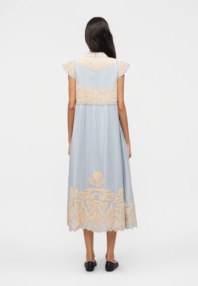 Γυναίκα με μακριά σκούρα μαλλιά που φοράει ανοιχτό μπλε μίντι φόρεμα με μπεζ κεντήματα λουλουδιών στην πλάτη και στο κάτω μέρος, συνδυασμένο με μαύρες μπαλαρίνες.