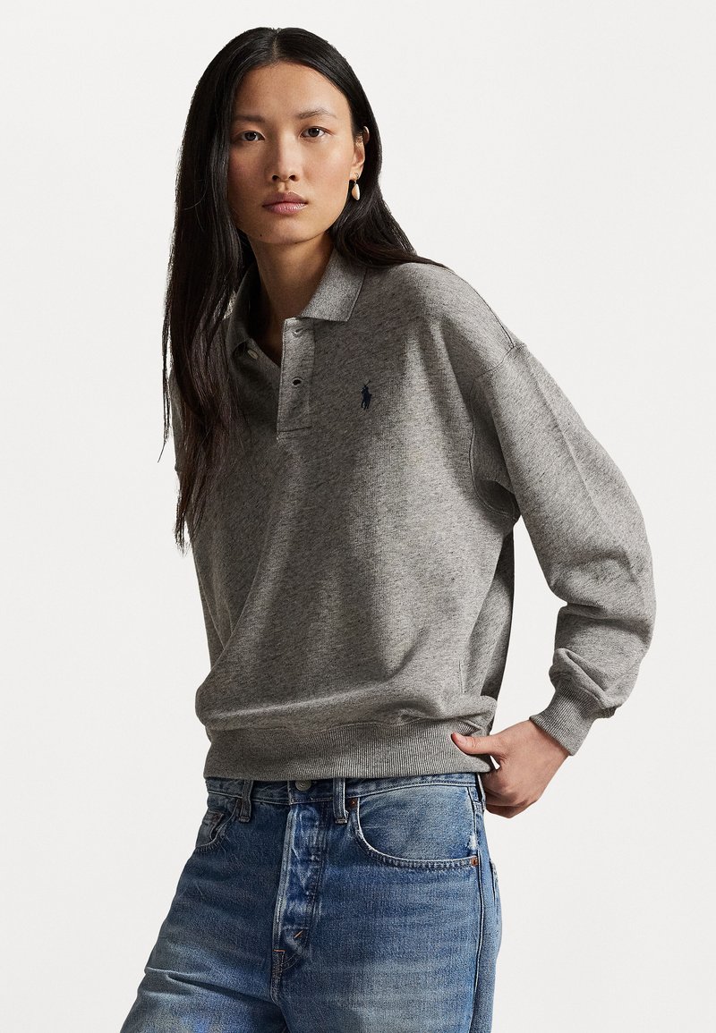 Halläärne polo-sweater vabamas lõikes, ribidega varrukate ja äärega, millel on rinnal väike tumesinine logo, koos siniste teksadega.