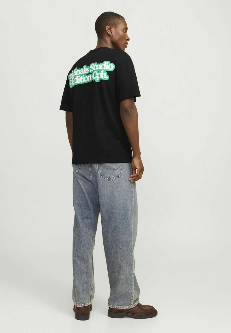 Camiseta negra de algodón con un ajuste oversize, que presenta texto verde en la parte posterior. Combinada con jeans baggy de lavado claro y botas marrones.