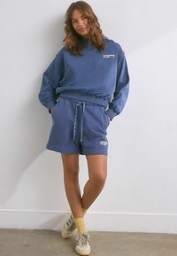 Completo in cotone blu composto da felpa e pantaloni shorts, con scollo a girocollo, vita elastica e grafica "RUNNERS CLUB" in bianco. Sneakers con accenti chiari.