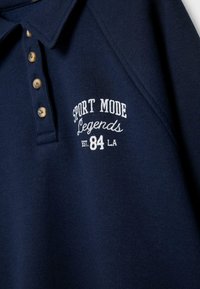 Navyblaues Polohemd mit beigen Knöpfen und weiß gesticktem Text "SPORT MODE Legends EST. 84 LA" auf der Brust.