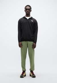 Μαύρο μπουφάν με κουκούλα, πράσινο παντελόνι και μαύρα και κίτρινα παπούτσια πεζοπορίας. Το μοντέλο στέκεται όρθιο, φοράει γυαλιά ηλίου, με απλό φόντο.