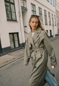 Kobieta spacerująca po ulicy miasta w zielonym płaszczu przeciwdeszczowym, z okularami przeciwsłonecznymi na głowie, trzymająca zieloną butelkę, z zamkniętymi oczami.