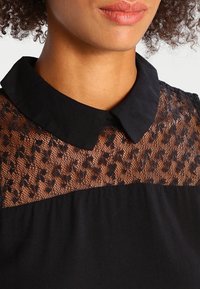 Robe noire avec un haut en dentelle transparente à motif floral, un col classique et un corsage ajusté se transformant en une partie inférieure lisse.