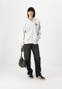 Harmaa vetoketjuhuppari, jossa on huppu ja etutasku, jossa on vihreä logo-teksti. Yhdistetty mustiin farkkuihin ja mustiin lenkkareihin, joissa on valkoiset raidat.