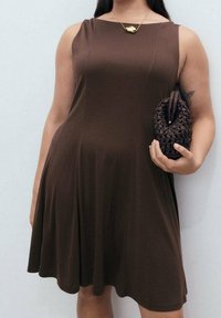 Robe marron sans manches en tissu doux, avec une jupe évasée. Le mannequin tient une pochette tissée foncée et porte un collier pendentif en or.