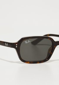 Ochelari de soare cu rame pătrate din tortoise shell, lentile întunecate, logo-ul argintiu Ray-Ban pe braț și două nituri decorative de fiecare parte.