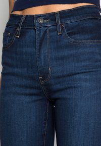 Jeans de denim azul-escuro com cintura alta, costura visível em ouro contrastante e fecho frontal com botão. Design clássico de cinco bolsos.