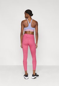 Μωβ αθλητικό σουτιέν με κόκκινο λογότυπο "Nike Pro", σετ με ροζ κολάν που έχει διακριτή υφή, φορεμένα με μαύρα και λευκά αθλητικά παπούτσια.