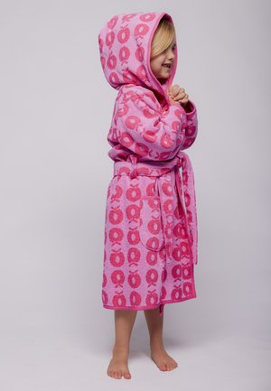 Ungt barn med blondt hår iført en lyserød badekåbe med hætte og røde æblemønstre, stående barfodet med sammenfoldede hænder.
