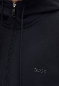 Svart hoodie med dragkedja, tillverkad av mjukt material. Har en strukturerad logotyp "BOSS" på bröstet. Tunna snören vid huvan.