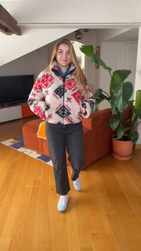 Νέα γυναίκα που φοράει κόκκινο, λευκό και μαύρο ζακέτα με σχέδιο, μαύρο παντελόνι και μπλε αθλητικά παπούτσια, στέκεται σε ένα σαλόνι με ξύλινο πάτωμα και φυτά.