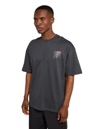 Młody czarnoskóry mężczyzna w luźnej, ciemnoszarej koszulce z małym graficznym logo na lewej piersi, stojący na białym tle.