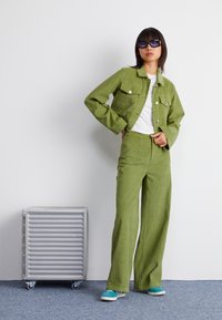 Chaqueta y pantalones de pana verde combinados con una camisa blanca y zapatillas en color verde azulado. El modelo está de pie junto a un armario de cajones plateado con ruedas.